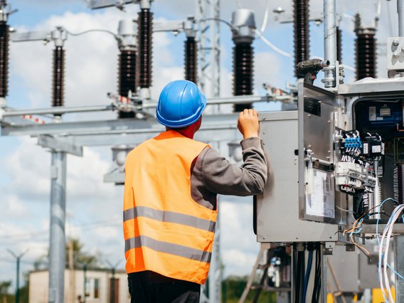 Person, die einen blauen Schutzhelm und eine orangefarbene Warnweste trägt und an einem offenen Schaltpult in einem Umspannwerk arbeitet. Im Hintergrund sind elektrische Anlagen und Hochspannungsleitungen zu sehen.