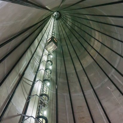 Aufzug_Windenergieanlage Das Innere eines großen, zylindrischen Behälters oder Silos. Von unten blickt man nach oben, wo eine Leiter mit Sicherheitskäfig und Beleuchtung bis zur Öffnung an der Spitze führt.
