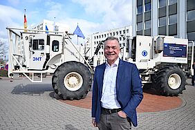 Ein Mann in einem blauen Blazer steht vor einem großen weißen Erkundungsfahrzeug (Vibrotuck)