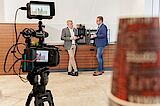 Was für uns spricht, sind unsere Mitarbeitenden Eine Videokamera filmt 2 Geschäftsleute beim Kaffeetrinken
